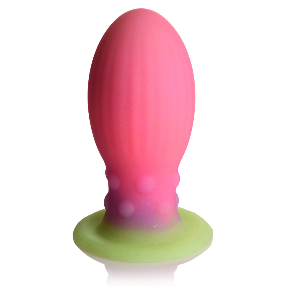 A glow-in-the-dark pink and green alien egg plug sex toy base rests on its suction cup against a white backdrop.