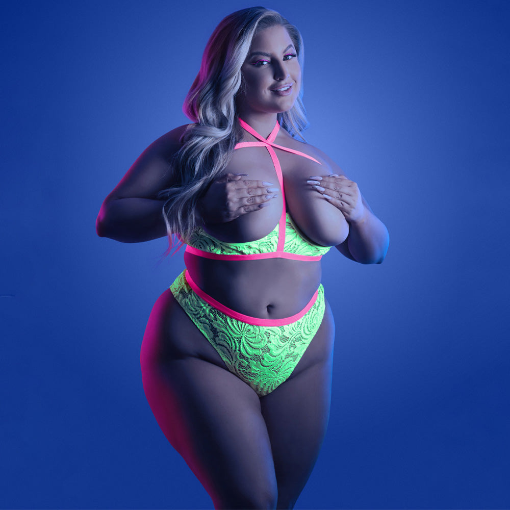 A plus-size model wears a glow-in-the-dark floral lace cupless bra and panty set with a cage strap halter neck design.