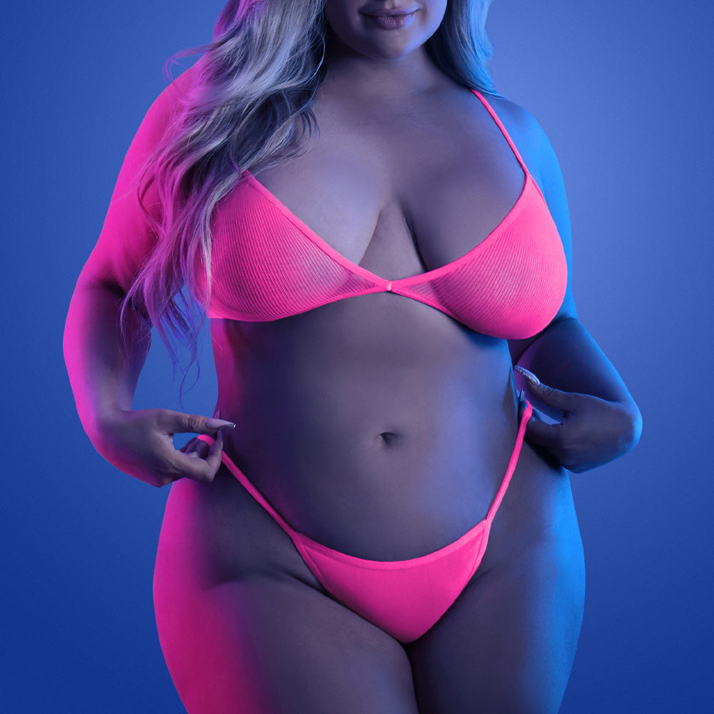 A plus size model wears a neon pink bralette with matching thong in a glow in the dark fishnet design.