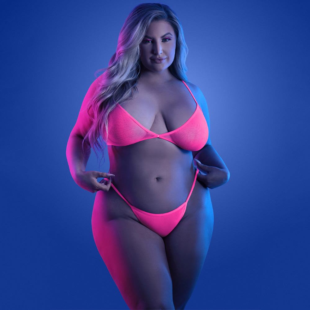 A plus-size model stands against a black light background wearing a neon pink fishnet bralette with high-waisted G-string.