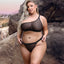 A plus-size model wears a one-shoulder crop top bra and panty in sheer black mesh with an adjustable shoulder strap.