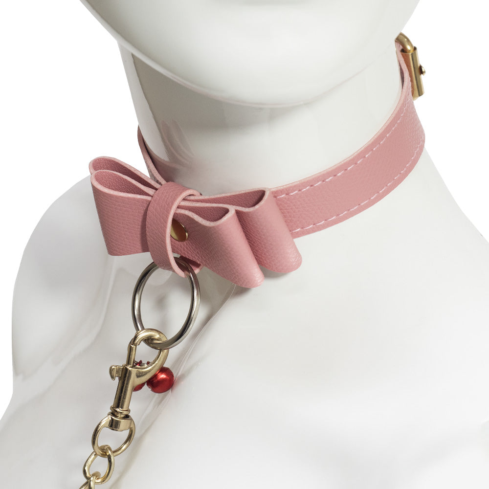 A mannequin wears a faux pink leather bow collar with red bells and a gold chain leash attached to a silver O-ring.