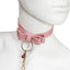 A close up of a mannequin's neck wearing a pink faux leather bowtie collar that features a silver O-ring and red bells.