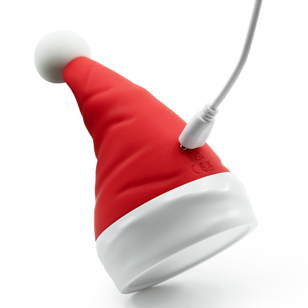 A red Santa hat-shaped clitoral vibrator leans on an angle to show its magnetic charging points and attached charging cord.