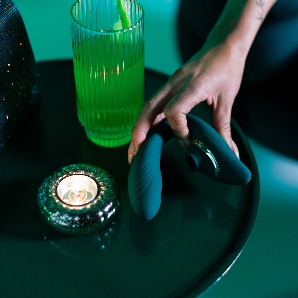 A models hand is holding a Sexyland Pamper G spot clitoral suction stimulator on a table next to a candle and drink.