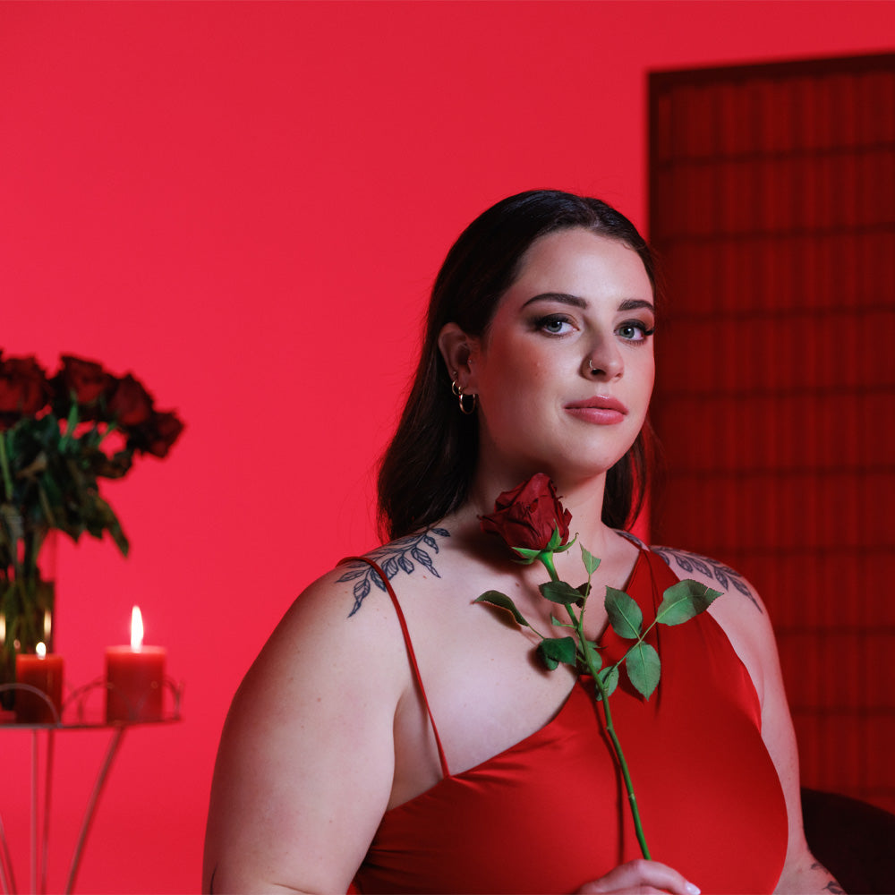 A model is holding a red rose and smiles at the camera with a candle and roses in the back against a red backdrop. 