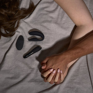 A couple holds hands with intertwined fingers in bed while We-Vibe's Tango X bullet and Sync 2 vibrators lay between them.