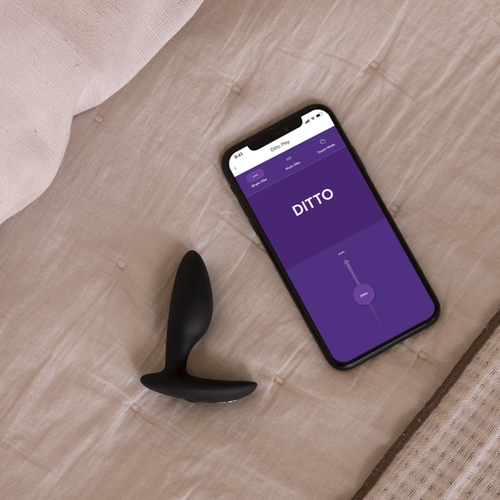 Editorial image of the app-compatible We-Vibe Ditto+ connecting to a smartphone displaying the We-Vibe app on its screen.