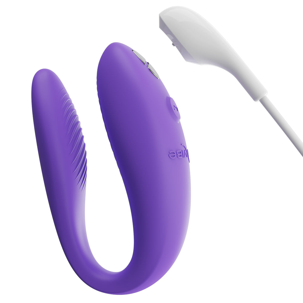 The We-Vibe Sync Go Couples Vibrator stands next to its magnetic charging cable against a white background.